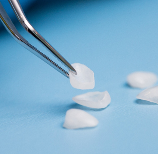 Closeup of tweezers grasping one of several veneers on a light blue surface